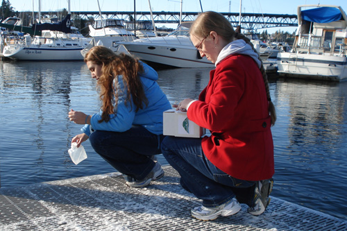 
                    Volunteer scientists Liana Singh and Brittany Kimball demonstrate how to gather a water sample.
                                            (Joshua McNichols)
                                        