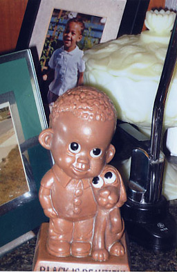 
                    Joe Wenderoth and Romana Norton's "Black is Beautiful" statue on the counter, among their family pictures.  This object is from the 1970s, and Romana sees it as a response to the racist and caricatured memorabilia of earlier times.  She says most of the black families she knew grewing up had a statue like this in their home.
                                            (Krissy Clark)
                                        