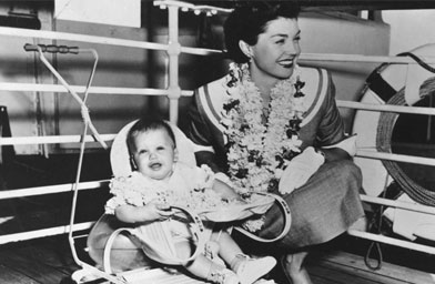 
                    Esther Williams, wearing a lei, on a ship's deck with her child, undated.
                                            (The Margaret Herrick Library)
                                        